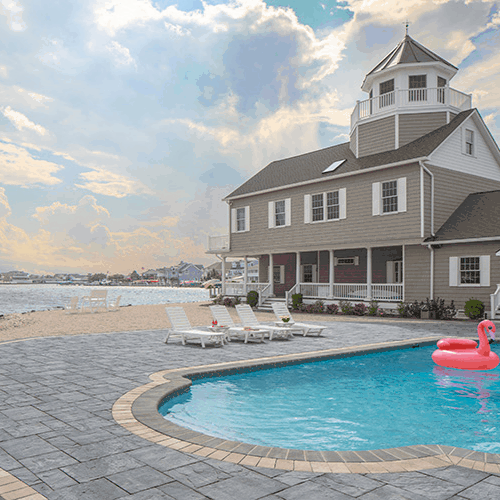 A scenic beach house with a tower overlooks a swimming pool decorated with a pink flamingo float. Lounge chairs on the spacious patio sit atop stylish paving stones from Masonry Supplies Queens County, with the beach and ocean visible in the background under a partly cloudy sky.