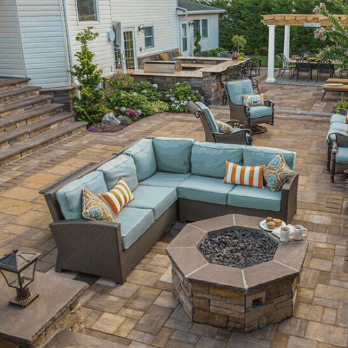 A spacious patio featuring a cozy L-shaped outdoor sofa with blue cushions and striped throw pillows. A hexagonal stone fire pit with snacks and drinks is in front of the sofa, crafted using top-quality masonry supplies from Nassau County. The area boasts various other seating arrangements, stairs, and vibrant greenery.
