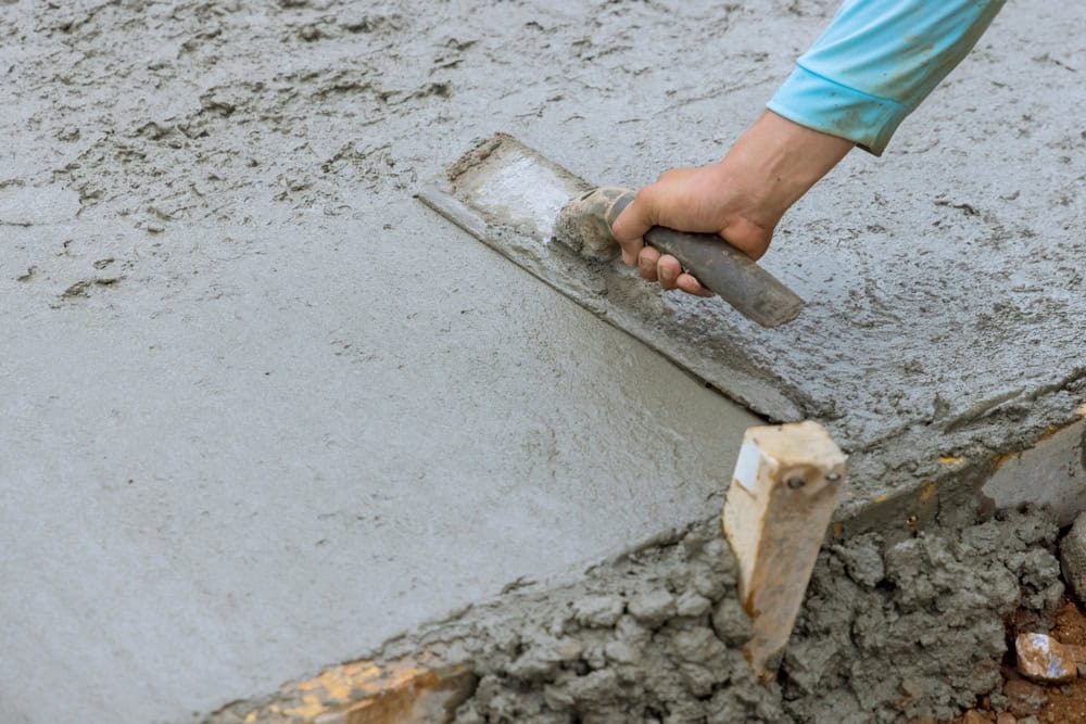 A person wearing a blue long-sleeve shirt uses a trowel to smooth out the surface of a fresh concrete slab, ensuring an even finish. The concrete appears wet and in the process of being spread and leveled. Some wooden supports are visible at the edge, likely sourced from masonry supplies Nassau County.